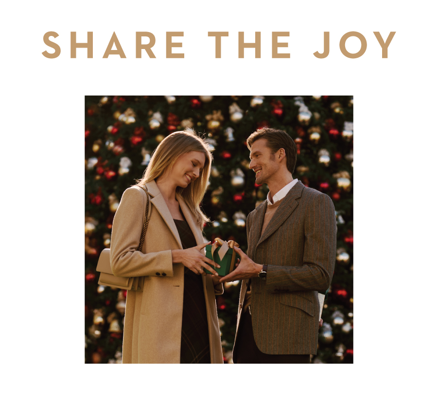 "Share the Joy" Text with a couple sharing a gift in front of a Christmas Tree